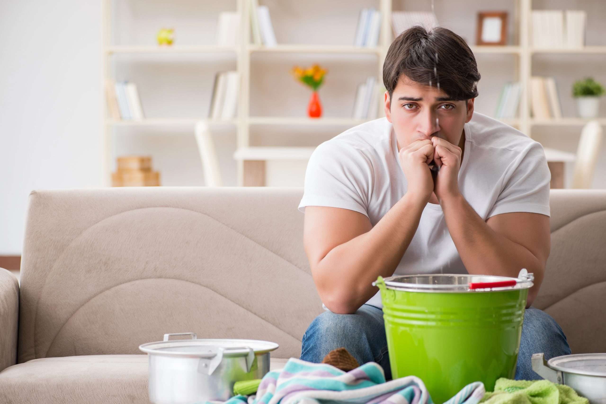 Worried man sitting on a beige couch in his living room, staring anxiously at a ceiling leak while a green bucket, pots, and towels on the floor catch dripping water.
