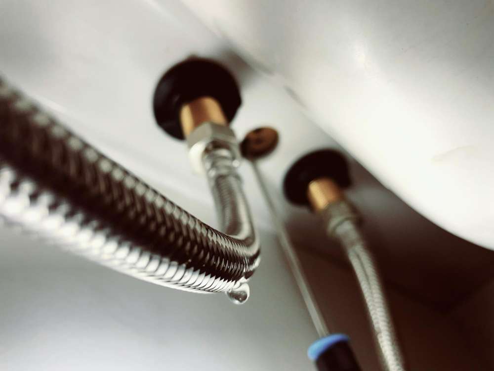 Close-up of a water heater’s braided supply lines with a visible drip forming underneath, indicating a small plumbing leak.
